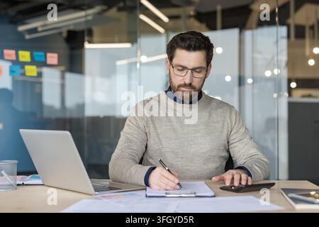 A Focused Businessman Working On A Laptop And Writing Documents For Financial Analysis At A Desk In A Corporate Office. The Professional Manager Is Carefully Reviewing Paperwork And Using. Stock Photo