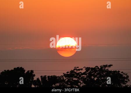 View of the sun setting behind clouds and power lines above tree silhouettes in Dhaka, Dhaka Division, Bangladesh. Stock Photo