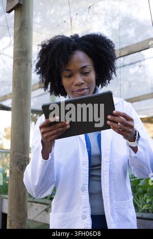 African woman wearing white lab coat holding black tablet while checking plants in greenhouse Stock Photo