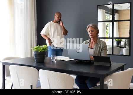 Diverse couple working at home dining senior woman using laptop, man talking on phone, copy space Stock Photo
