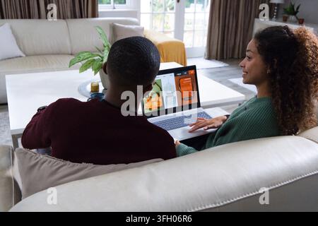 Couple sitting on sectional sofa at home, browsing shopping site on silver laptop near coffee table Stock Photo