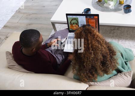 Diverse couple sitting on beige sofa, browsing online store on laptop, holding credit card Stock Photo