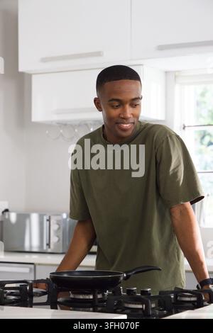 African American adult man wearing olive T-shirt, standing at gas stove, tending black frying pan Stock Photo