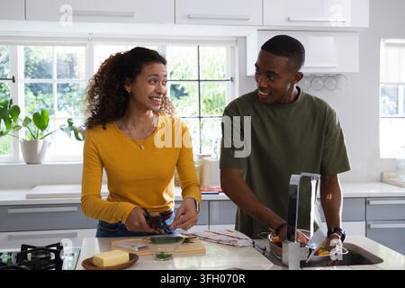 Diverse couple cutting vegetables with scissors at home kitchen island while rinsing in colander Stock Photo