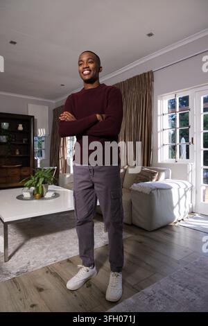 Man standing with arms crossed, smiling, wearing maroon sweater and gray cargo pants in living room Stock Photo