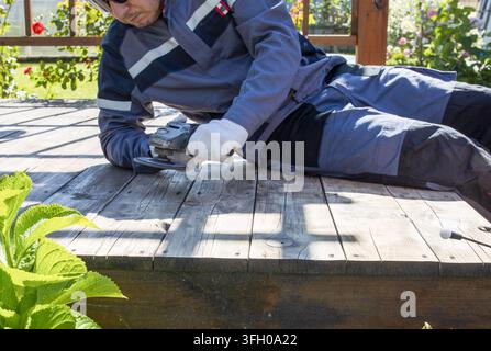 Man lying on wooden deck, using angle grinder. Dust flies as he works. Sunlight casts shadows across surface Stock Photo