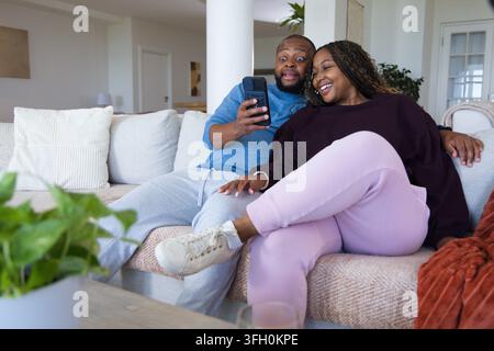 African couple sitting on light sectional sofa at home, man holding smartphone, woman smiling Stock Photo