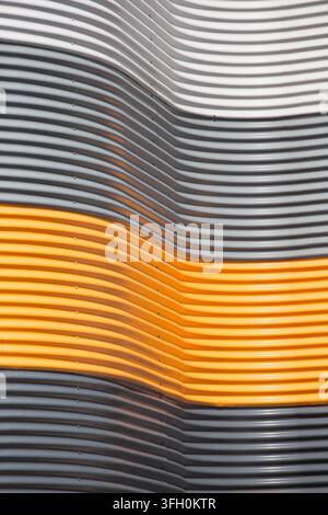 Close-up of undulating white, grey, and orange corrugated metal panels on a modern building facade, creating abstract patterns and texture. Stock Photo