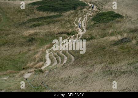 Hikers trek up a winding terraced path through coastal grassland at Seaford Head on the South Downs Way, East Sussex, UK. Stock Photo