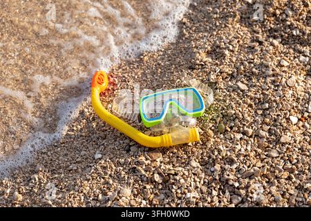 A snorkeling mask and snorkel lie on a pebbled beach beside foamy surf. Stock Photo