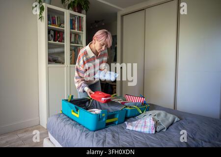 Focused woman preparing for holiday trip and attentively packing suitcase on bed in bedroom. Stock Photo