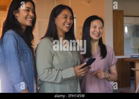Three female friends smiling and leaning in, center woman holding smartphone on island in kitchen Stock Photo