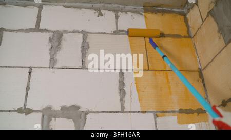 Construction worker applying orange waterproofing paint with a roller on a white brick wall, ensuring protection against moisture Stock Photo