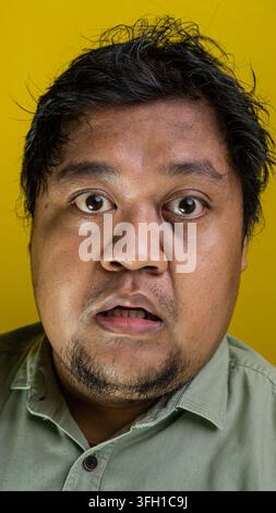 Striking frontal portrait showcasing a young Indonesian male displaying a deeply shocked and surprised facial expression against a vibrant yellow back Stock Photo