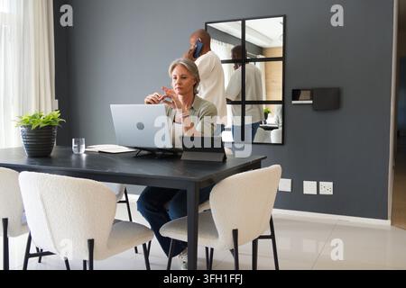 Diverse couple working at home dining table, woman using laptop on stand, man talking on smartphone Stock Photo