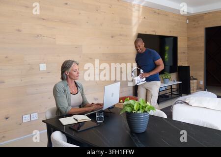 Diverse couple at home, woman working on laptop while man holding white VR headset, copy space Stock Photo