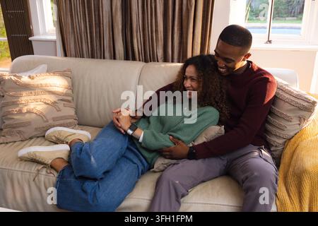 Diverse couple holding hands, laughing while woman lying on man's lap on leather sofa yellow throw Stock Photo