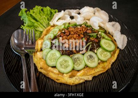 Tahu telur or fried tofu omelet topped with peanut sauce, cucumber slices, fried peanuts, crackers, and lettuce served on black plate. Stock Photo