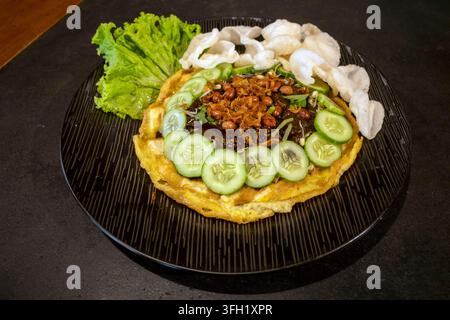 Tahu telur or fried tofu omelet topped with peanut sauce, cucumber slices, fried peanuts, crackers, and lettuce served on black plate. Stock Photo