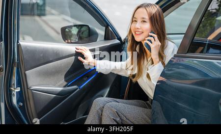 In the urban landscape, an Asian businesswoman stands beside her car, holding a phone and an open door. She exudes confidence and success as she manag Stock Photo