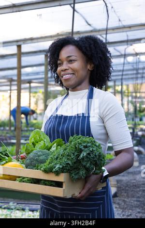 African woman holding crate of kale and broccoli in greenhouse wearing striped apron and watch Stock Photo