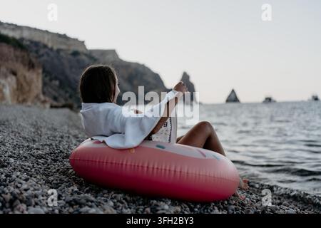 Woman, beach, sea. Young woman on inflatable ring relaxes on pebble beach, enjoying tranquil water, cliffs at dusk. Stock Photo