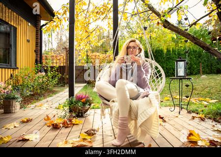 A mid adult woman enjoys a warm drink in a swinging macrame chair on a cozy autumn patio. Mental clarity outdoors during a mindful routine. Stock Photo
