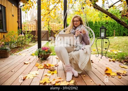 A mid adult woman works remotely on a laptop while drinking tea in a hanging swing chair on an autumn patio. Stock Photo