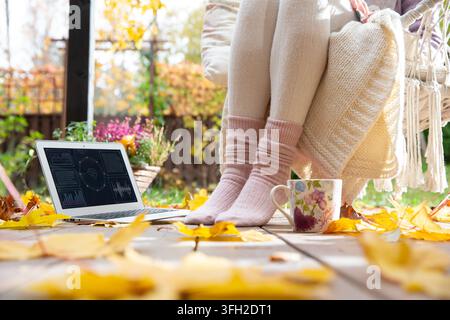 A mid adult woman relaxes on an autumn patio with a laptop mockup, blank screen, and coffee mug - cozy remote work break template concept. Stock Photo