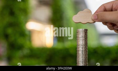 Cloud computing investment cost and digital technology funding concept with hand holding wooden cloud icon above stacked money coins in modern office Stock Photo