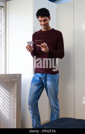 African man wearing maroon sweater holding credit card and using phone near window blinds at home Stock Photo