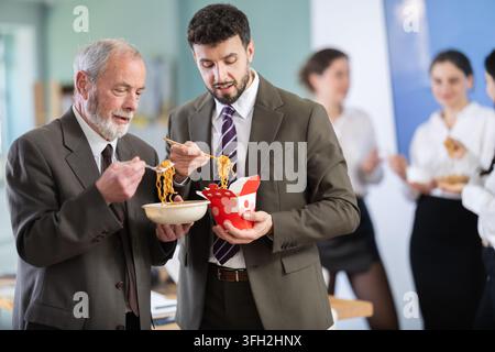 Positive two male office workers eating food together during lunch time in office of business company Stock Photo
