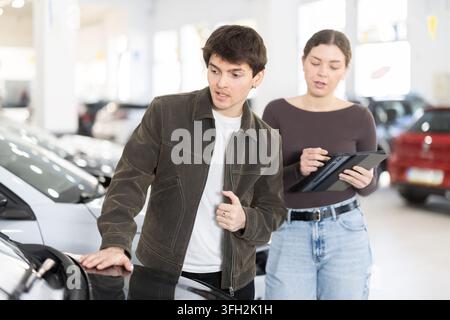 Young couple is standing in a car dealership and looking at the car with gadget in hands Stock Photo