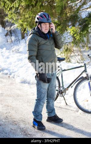 A senior man talks on his mobile phone beside his bicycle on an icy path in Helsinki, Finland, during winter commuting Stock Photo