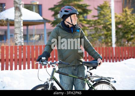 A senior man stands with his bicycle on an icy path in a snowy Helsinki park, Finland, enjoying healthy winter exercise. Stock Photo