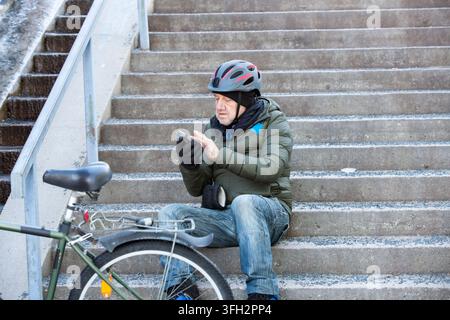 A senior man uses his smartphone while sitting on stairs beside his bicycle in Helsinki, Finland, resting during his winter commute. Stock Photo