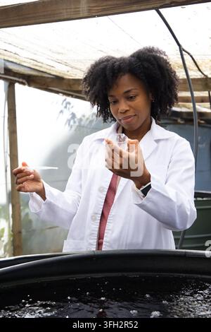 Woman in 30s researcher wearing lab coat, holding pipette and sample cup at black water tank Stock Photo