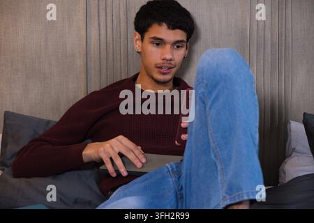 Male sitting on bed in maroon sweater and light blue jeans, holding smartphone, tablet Stock Photo