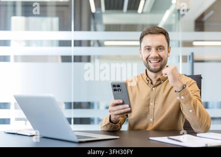 Business success and professional achievement, happy man celebrating with smartphone in modern office. Successful employee using mobile technology for communication and project management. Stock Photo