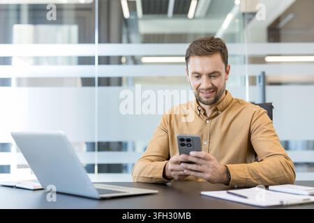 Digital communication and remote work, professional man using smartphone and laptop for business productivity in modern office. Corporate employee managing tasks with mobile technology and computer. Stock Photo