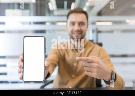 Mobile app promotion and digital marketing concept, man pointing at blank smartphone screen for mockup in office. Professional business communication and technology strategy. Stock Photo