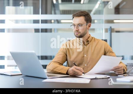 Productivity and remote work, professional man working on laptop and reviewing documents in modern office. Business strategy and data analysis, focused employee managing workflow and checking reports. Stock Photo