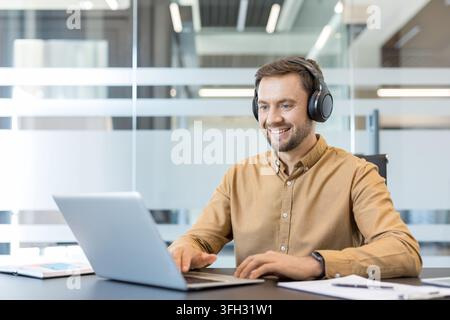 Remote work and digital communication, smiling man using laptop and headphones in modern office. Professional employee typing on computer and attending video conference, freelancer collaborating. Stock Photo