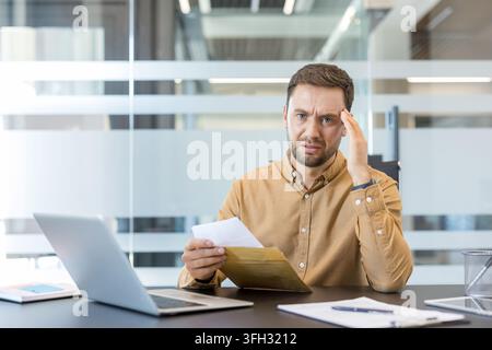 Work stress and financial anxiety, man reading bad news in a letter at office desk with laptop. Professional employee experiencing burnout and job insecurity while reviewing important documents. Stock Photo