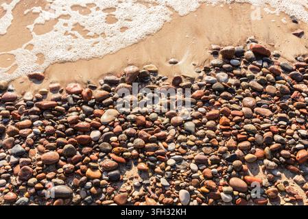 Colorful wet pebbles and sea foam on a sandy beach shore. Stock Photo