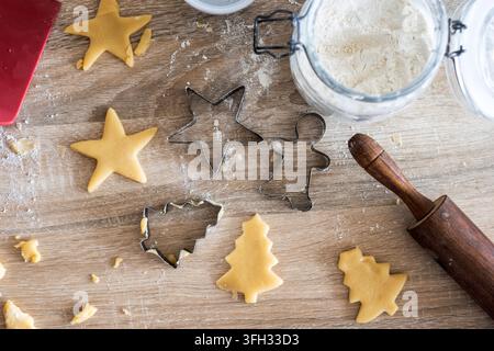 Raw Christmas cookie dough shaped into stars and trees with metal festive cookie cutters, rolling pin and flour on a wooden kitchen table. Stock Photo