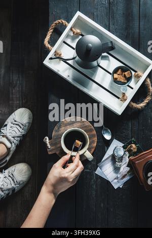 Flat lay of a hand putting a sugar cube into a cup of black coffee with a kettle and polaroids on a dark wooden background Stock Photo