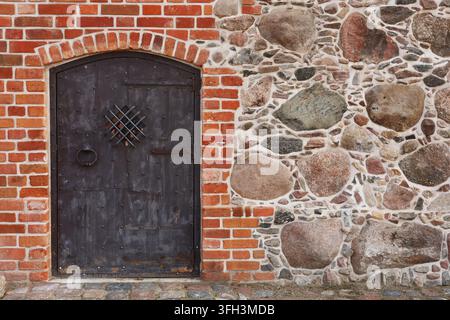 Weathered wooden door framed by red brick and rough stone wall. Dark metal handle and diamond-shaped grille add rustic charm. Texture contrasts emphas Stock Photo