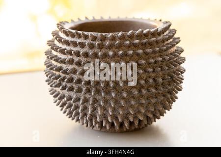 A dark, textured pot with a spiky surface sits on a light table Stock Photo