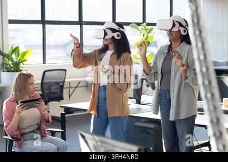 Three female coworkers using white VR headsets, gesturing while peer holding tablet in office Stock Photo